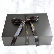Load image into Gallery viewer, A matte black gift box tied with a black satin ribbon printed with the brand name 'EKA' in gold, set against a white marble background.
