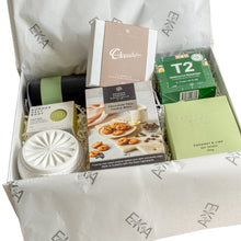 Load image into Gallery viewer, A curated gift box filled with assorted packaged snacks and treats, including a chocolate bar box, chocolate chip cookie, green tea, and a coconut soy candle, body wash, soap  bar, white cannister in a white magnetic gift box
