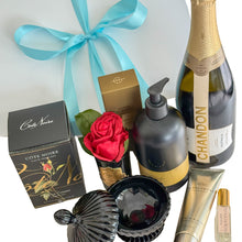 Load image into Gallery viewer, An open gift box filled with premium items, including a bottle of Chandon Brut, a black and gold body wash, Glasshouse Kyoto In Bloom hand cream, a black decorative cannister, and a Côte Noire red single rose bud in a black glass jar, all arranged on white tissue paper printed with the EKA logo
